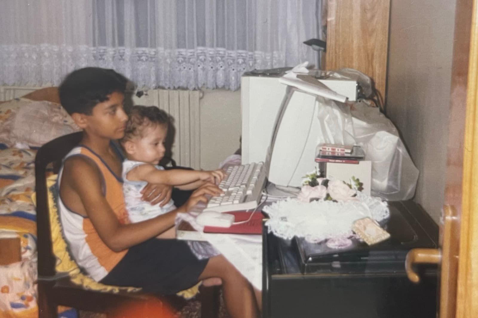 Okan Özalan de niño frente al ordenador de la familia, con su hermano Atakan, aún bebé, en el regazo; el bebé alarga la mano hacia el teclado de un ordenador beige con monitor CRT.