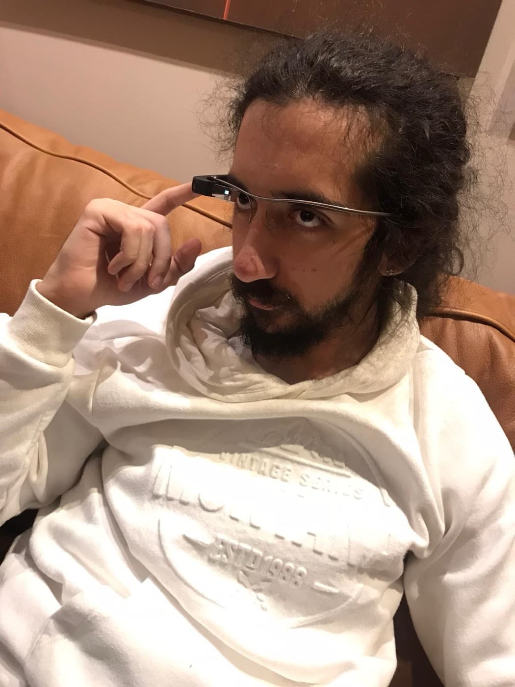 Atakan Özalan on a leather sofa in a white hoodie, wearing Google Glass and touching the side of the frame with one finger.